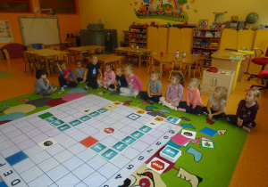 Grupa dzieci siedzi wokół maty do kodowania na której ułożone są dwie drogi z tabliczek kierunkowych aut do czerwonej i zielonej tabliczki.